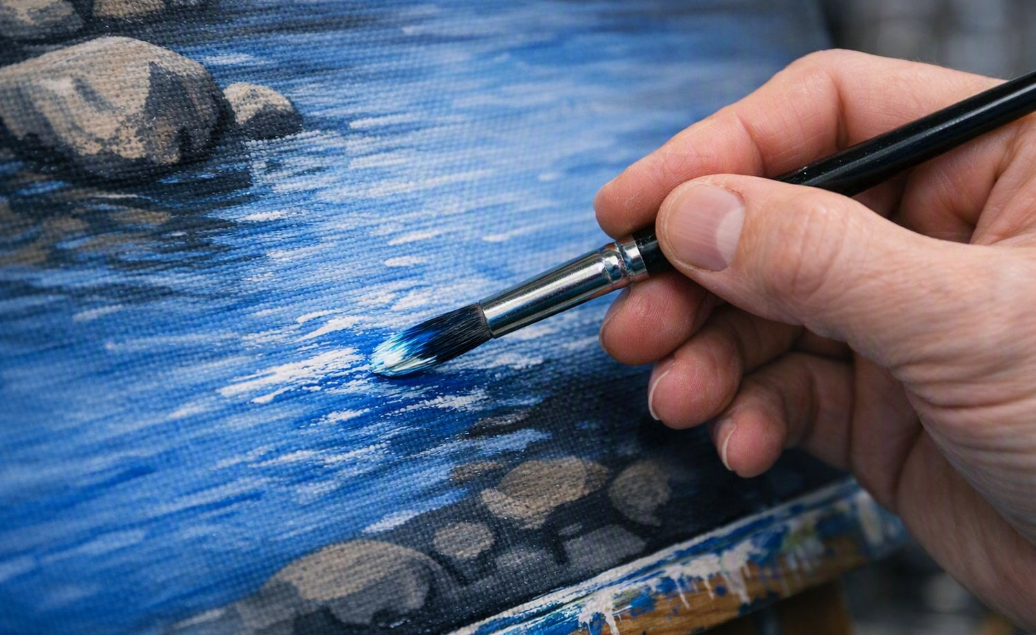 A close-up of a hand holding a paintbrush and painting a scenic landscape on canvas, showing blue water and gray rocks.