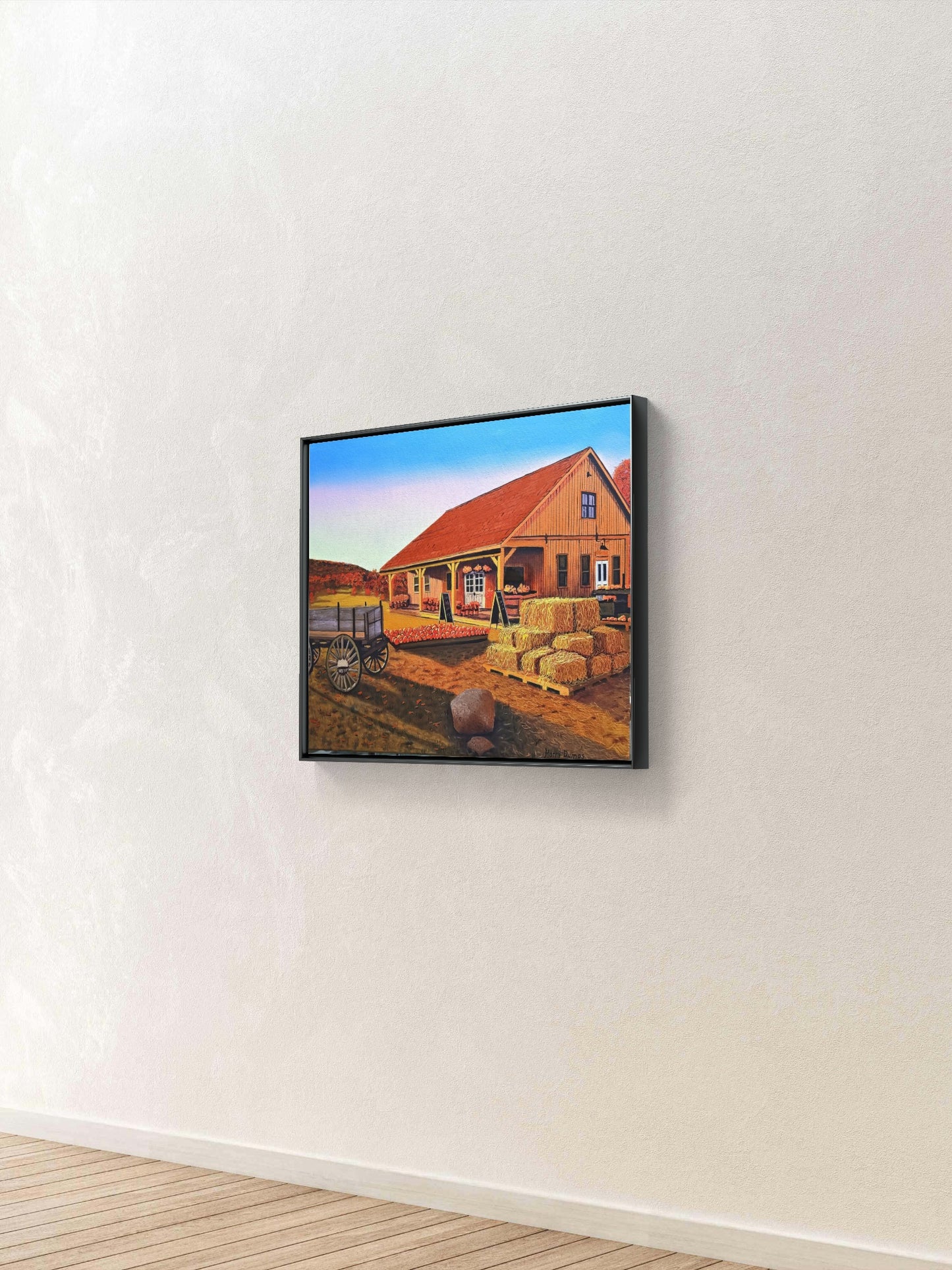 A framed landscape art print depicting a farm scene with a red-roofed house, hay bales, and a blue sky, displayed on a textured off-white wall above a light wood floor.