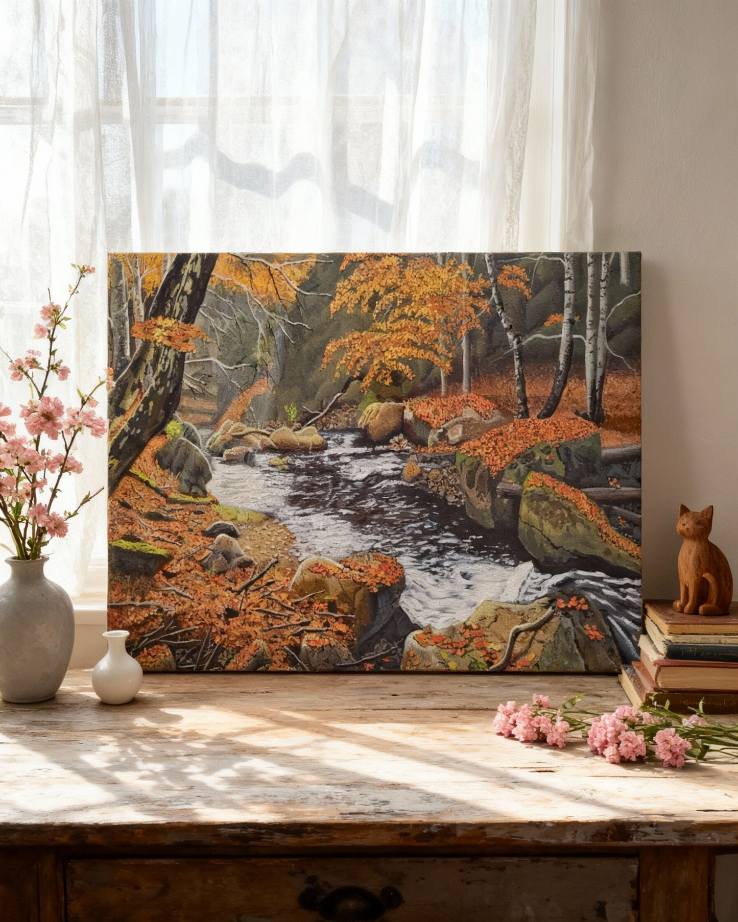 Painting on Canvas showing an autumn forest stream scene with orange foliage, rocks, and birch trees, displayed on a wooden table in a styled room with a vase, small figurine, books, and pink flowers. 
